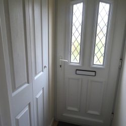 Modern white UPVC front door with panels, lattice windows / porch-cupboard Photo showing a modern white UPVC front door, with two long leaded lattice windows, a white door handle and letterbox, and rectangular door panels, which match the adjacent porch cupboard, where coats and shoes can be hidden away out of sight. A dark doormat and tiled porch floor can be seen at the base of the front door.